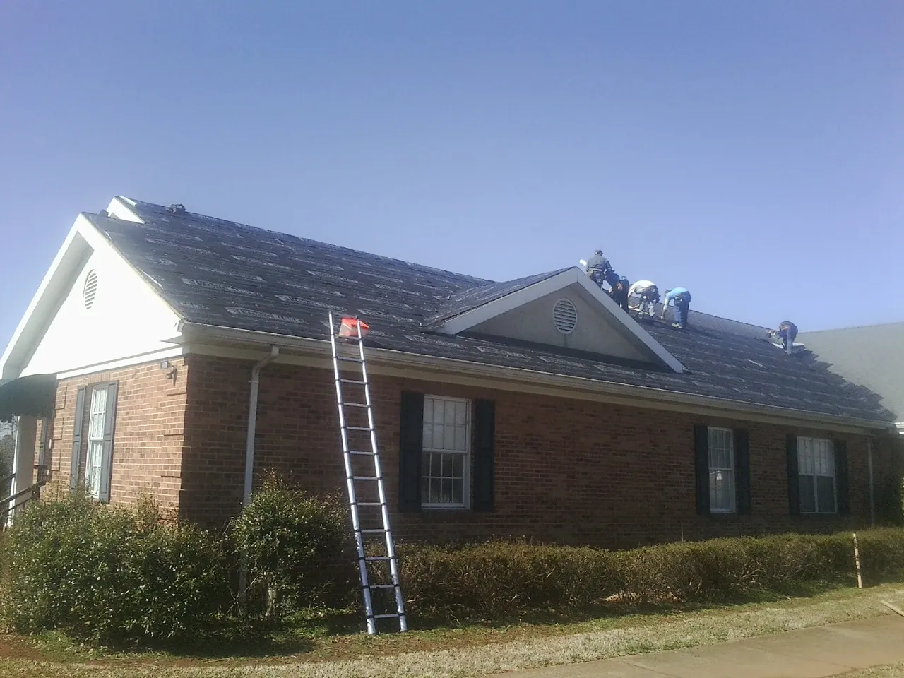 Crew tearing shingles off a brick ranch roof with a ladder set — Emergency Roof Repair work by our crew in Dunellen