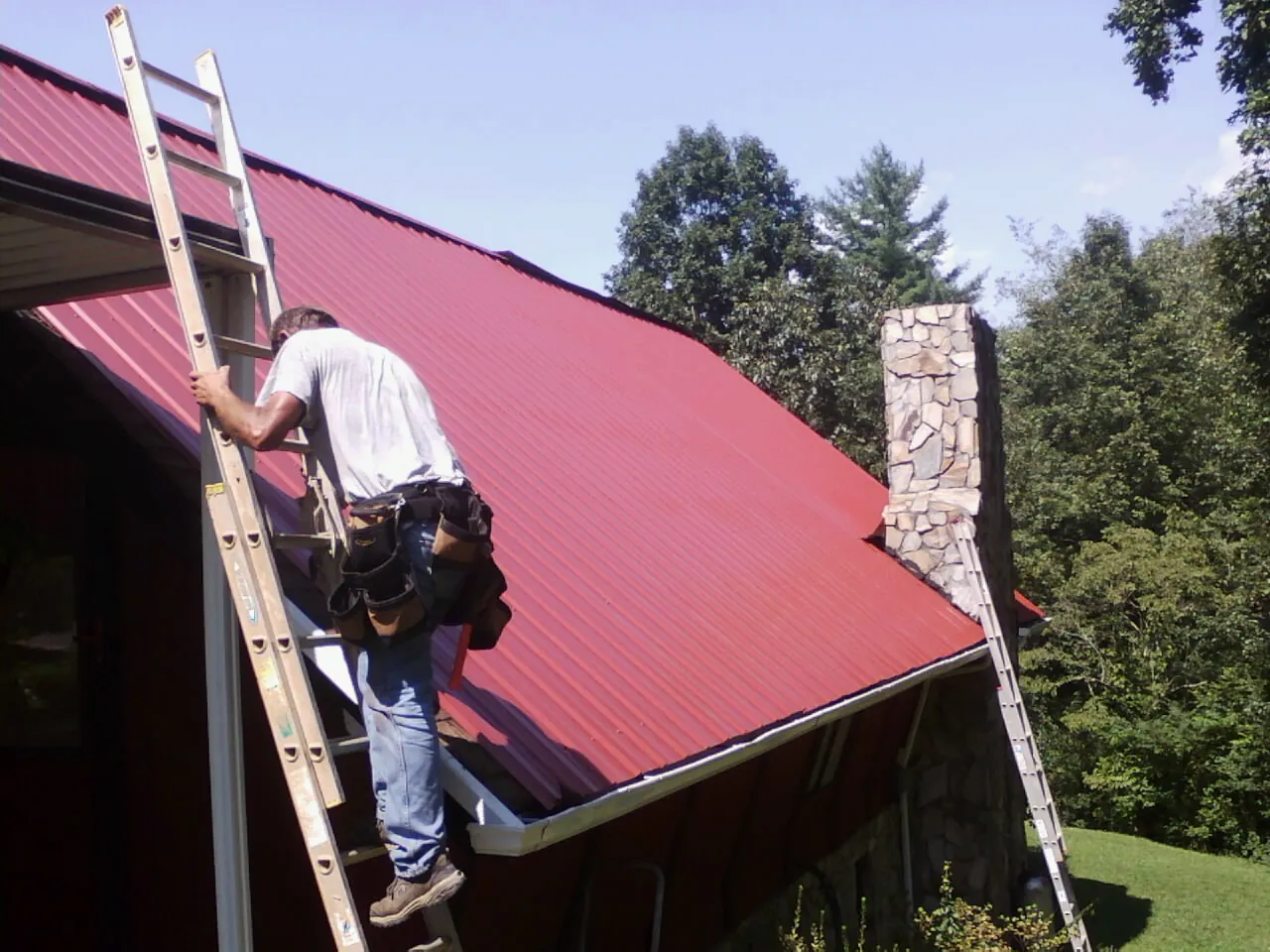 Roofer on a ladder beside a red metal roof and stone chimney — Soffit & Fascia Repair by Roofing Brothers in Dunellen